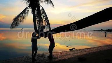 热带日落，棕榈树剪影在海边，幸福的家庭剪影与女儿在棕榈树上玩耍